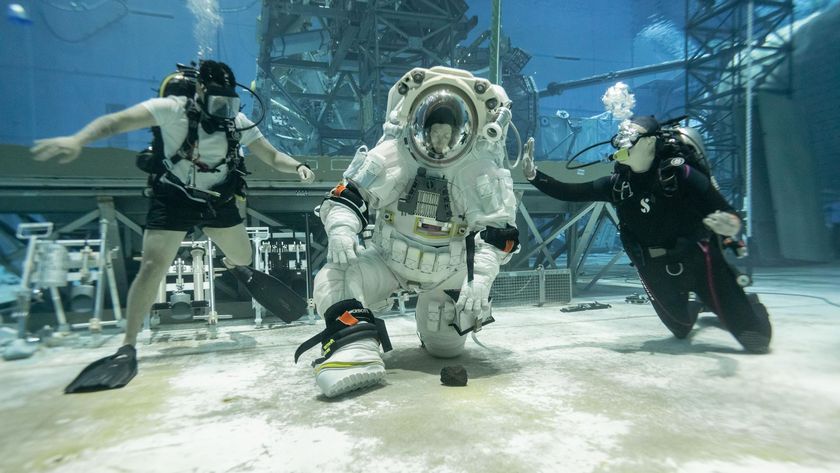 A person wearing a white spacesuit with a clear helmet kneels down to pick up a rock at the bottom of a large sandy pool with two people wearing dark swimsuits on either side of them