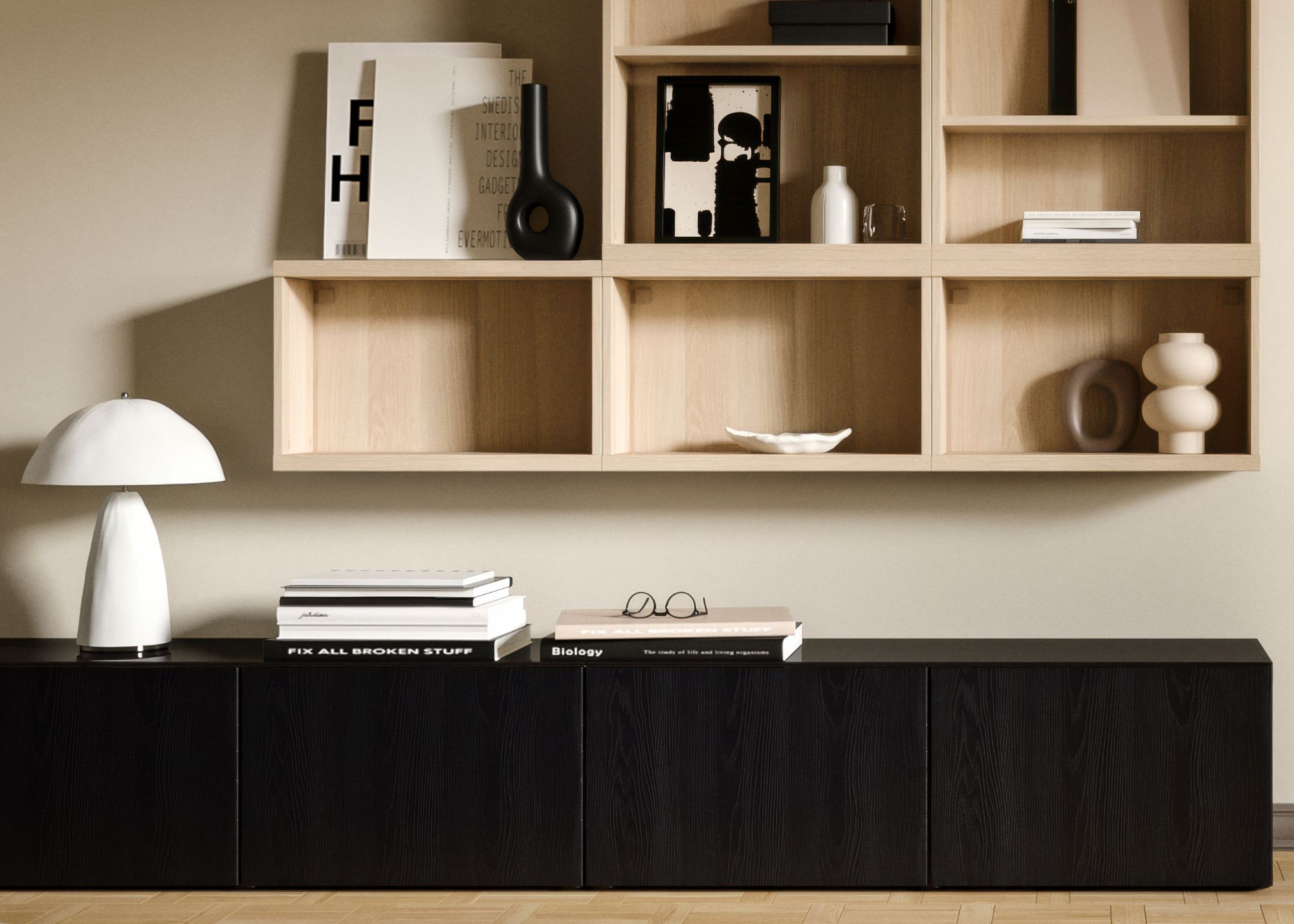 A neutral living room with a low black sideboard with a white table lamp and books on top underneath pale wood open shelving with decor objects