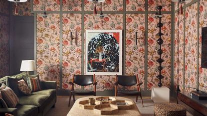 A maximalist living room with busy pink floral wallpaper, a green velvet sofa, two black leather and wood armchairs, and a light wood rectangular coffee table.