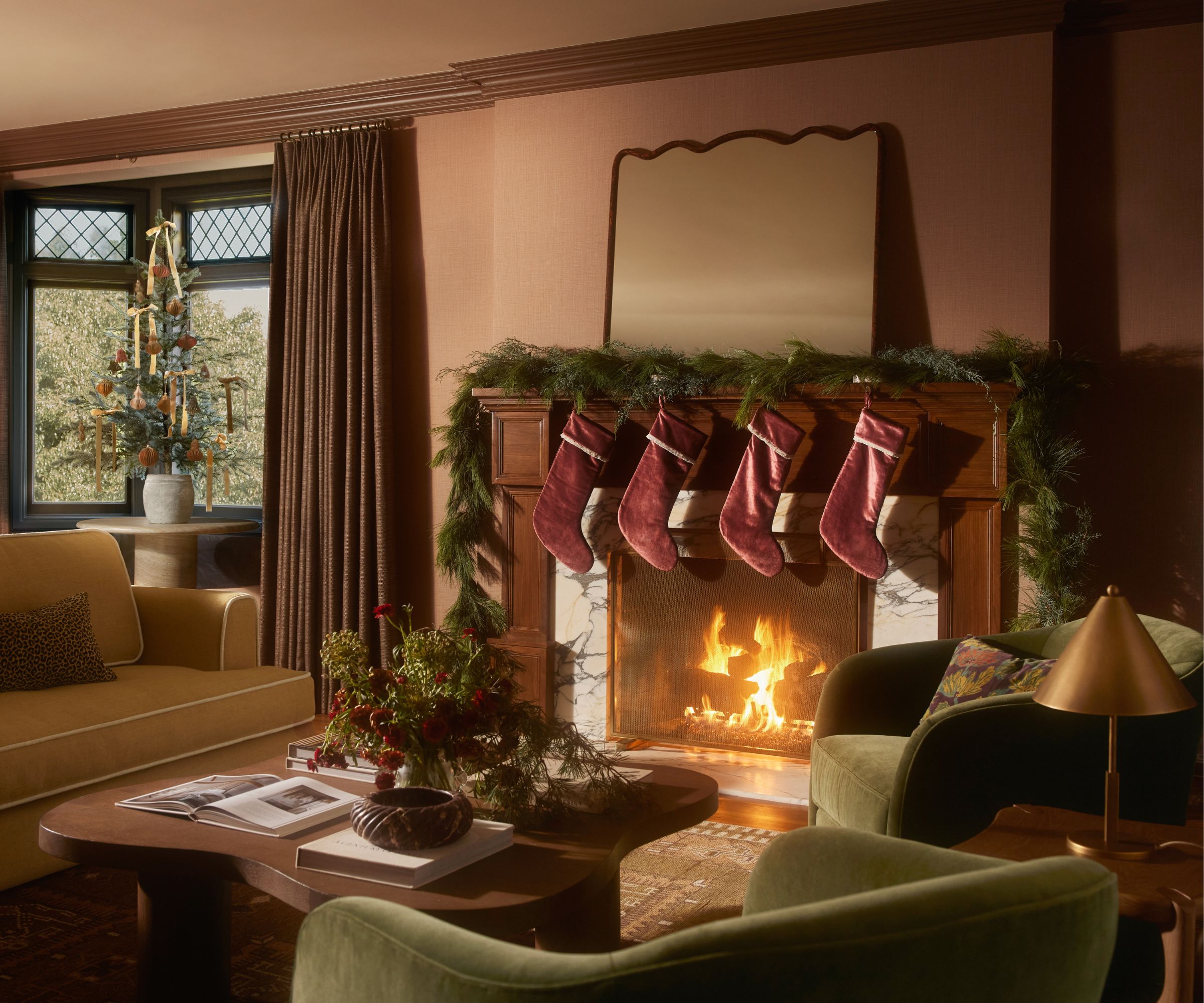 lulu and georgia christmas 2025 campaign imagery of a moody, warm living room with lit marble fireplace decorated with a garland, velvet stockings, a velvet couch, coffee table, green armchairs and a christmas tree