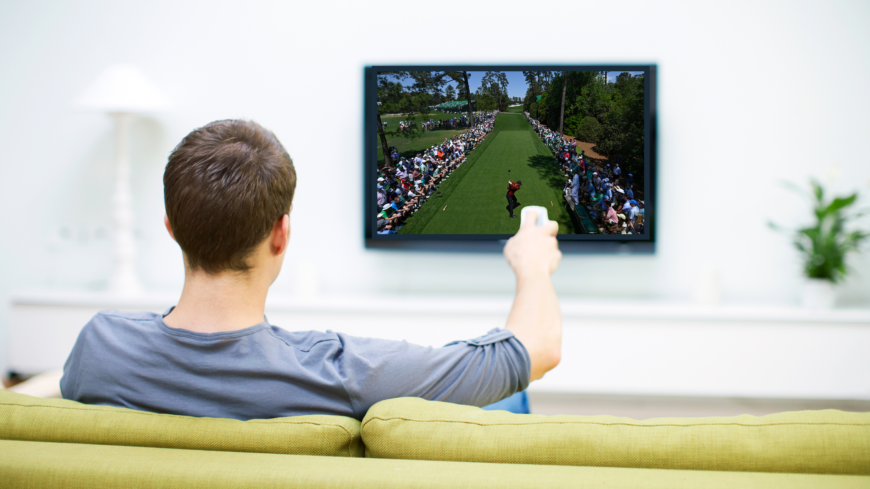 Man on his sofa pointing the remote at the television with an image of Tiger Woods hitting a tee shot at The Masters in front of the television screen