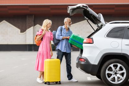 Happy caucasian senior couple going car trip together. Cheerful elderly man and woman in casual travelers getting ready before journey by auto, loading vehicle trunk with luggage, stand outdoors