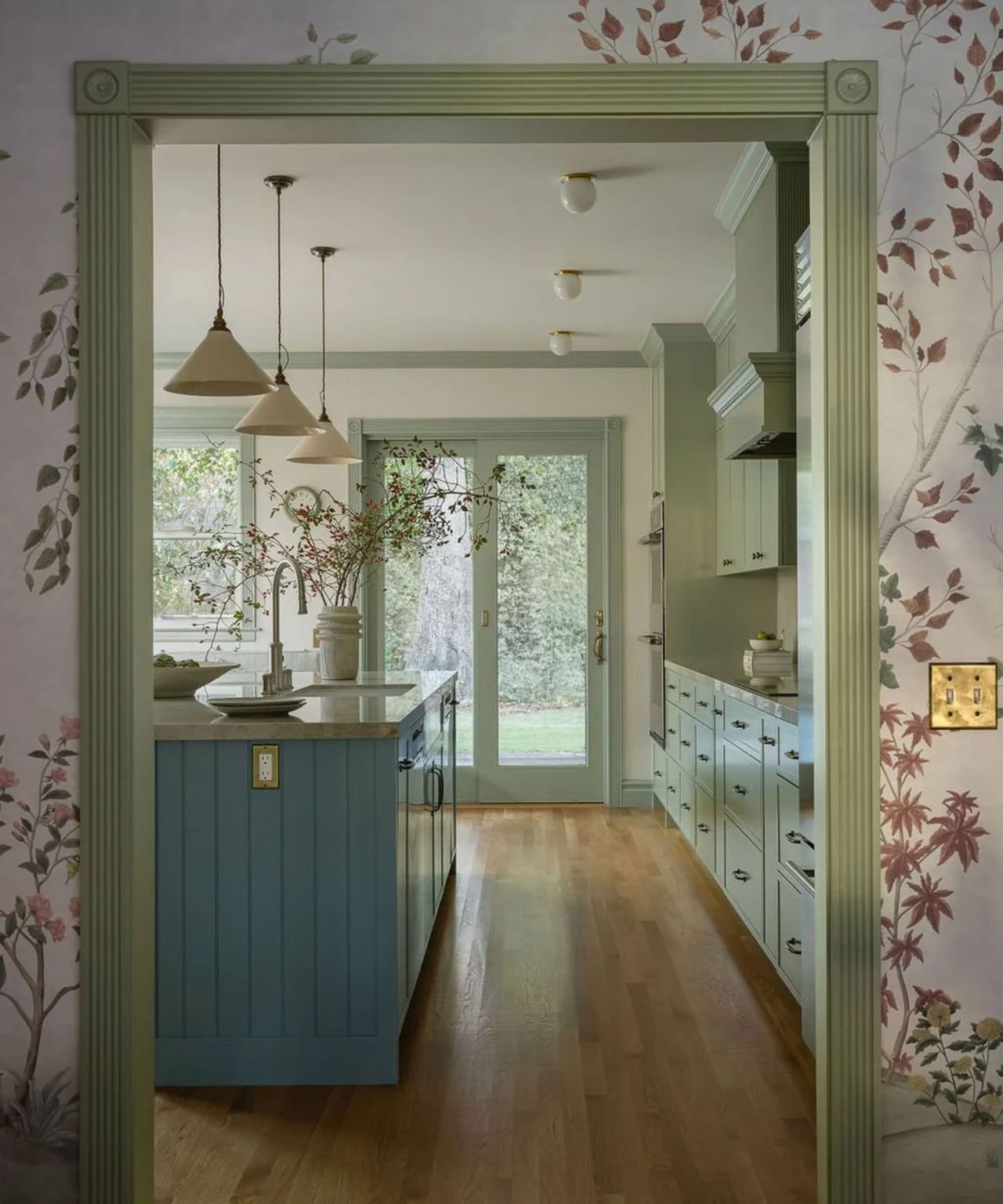 A view looking through a fluted sage green doorway into a kitchen. The foreground features floral wallpaper, leading the eye toward a blue kitchen island and wall-to-wall sage green cabinetry