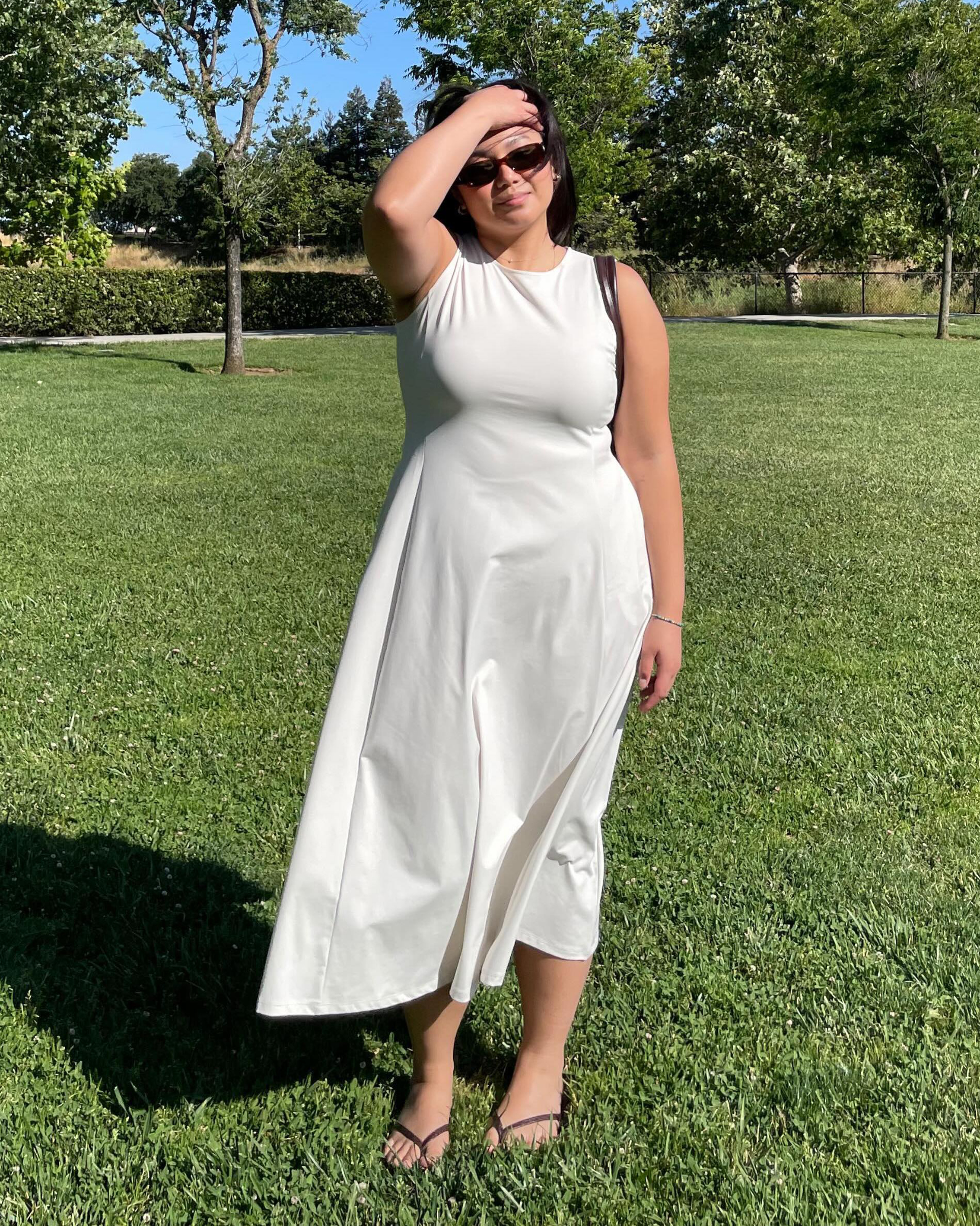 fashion influencer Marina Torres poses in a field of grass wearing a simple sleeveless white midi dress, sunglasses, and flip-flop sandals