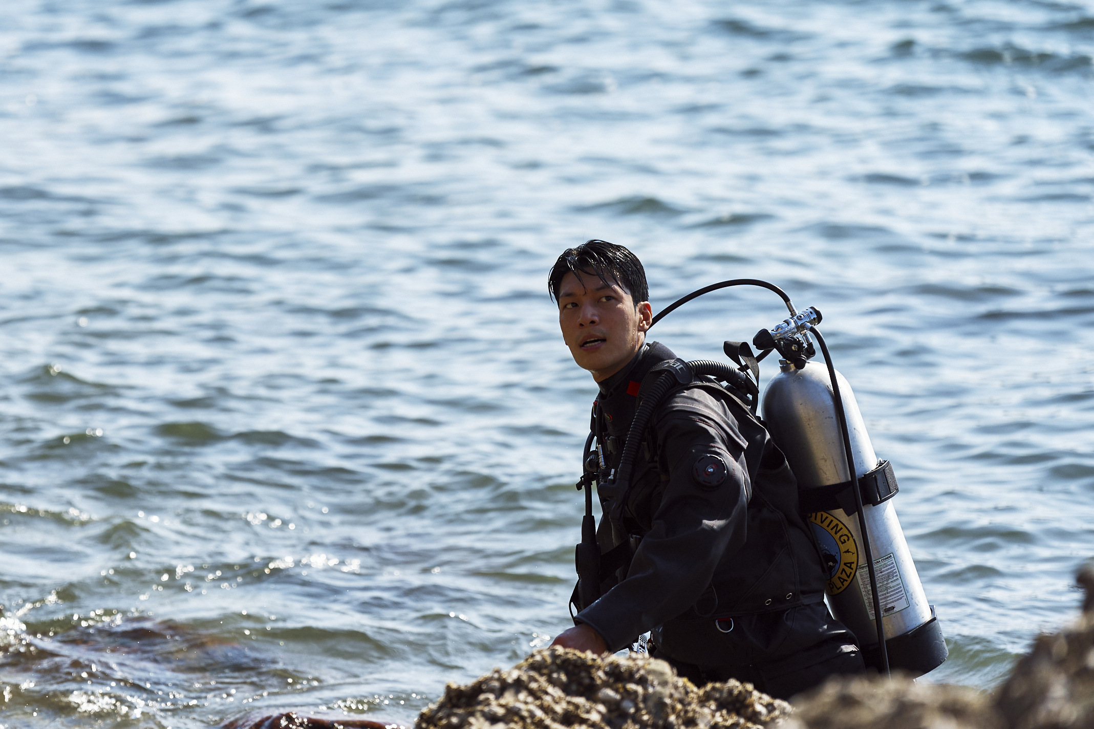A man (Wi Ha-jun as Jun-ho) in scuba gear with an oxygen tank, sits on the shore of an island, in 'Squid Game' season 2.