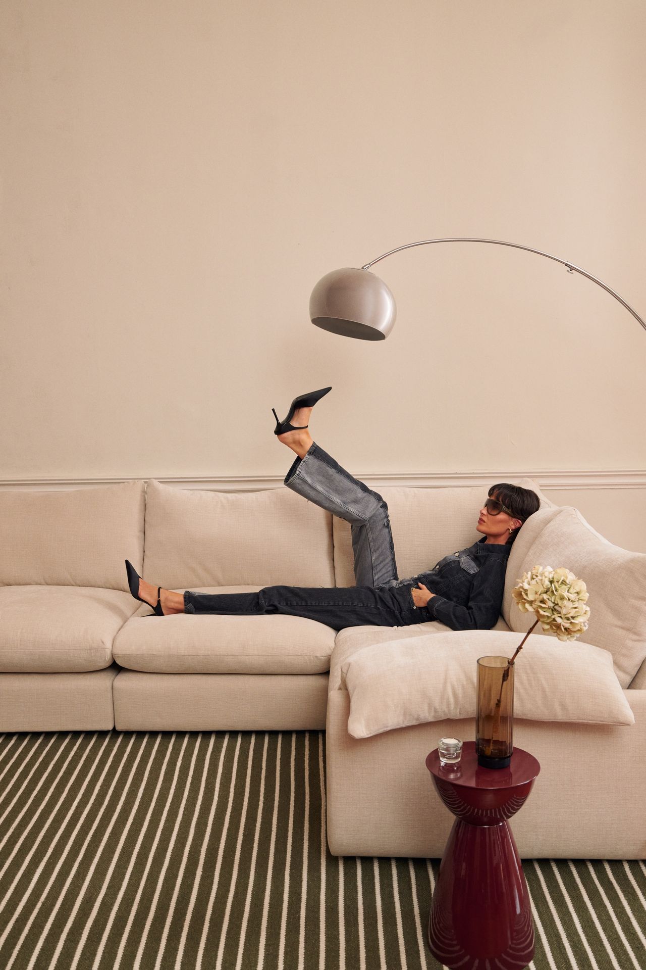 Image of a person in jeans and heels laying on a white sectional sofa with one leg in the air. There is a green striped rug, a burgundy side table, and a silver floor lamp in the room.
