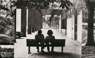 Black and white image of a couple sitting on a bench which has A+B written on the back