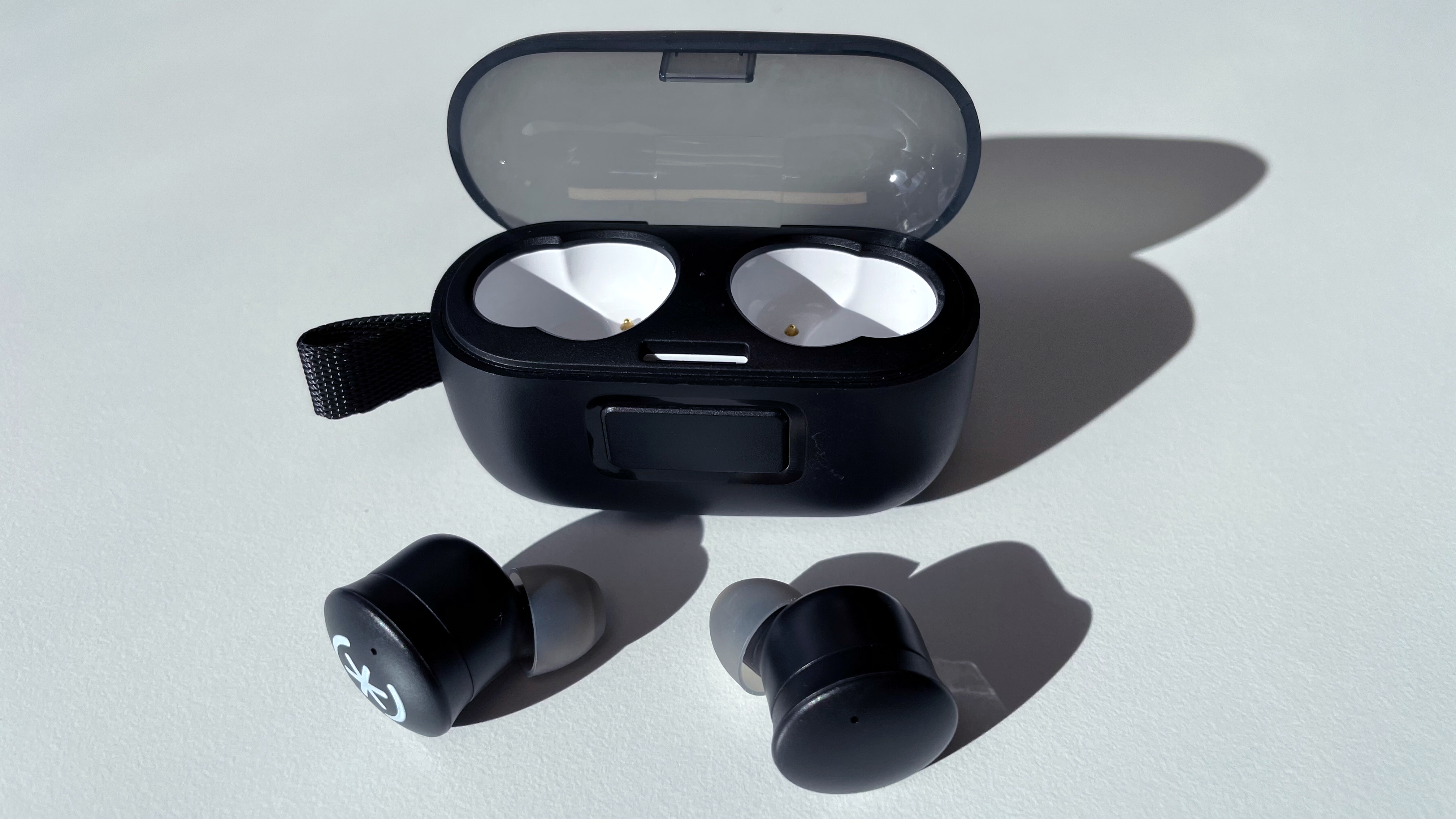 The speck Gemtones Play earbuds are pictured lying on a white tabletop in front of their charging case. The ear buds are black, with the left one having the Speck logo printed in white. The case is also black, with white inside the earbud cavities.