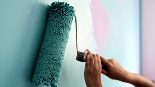 Close of roller with teal paint painting over pink wall