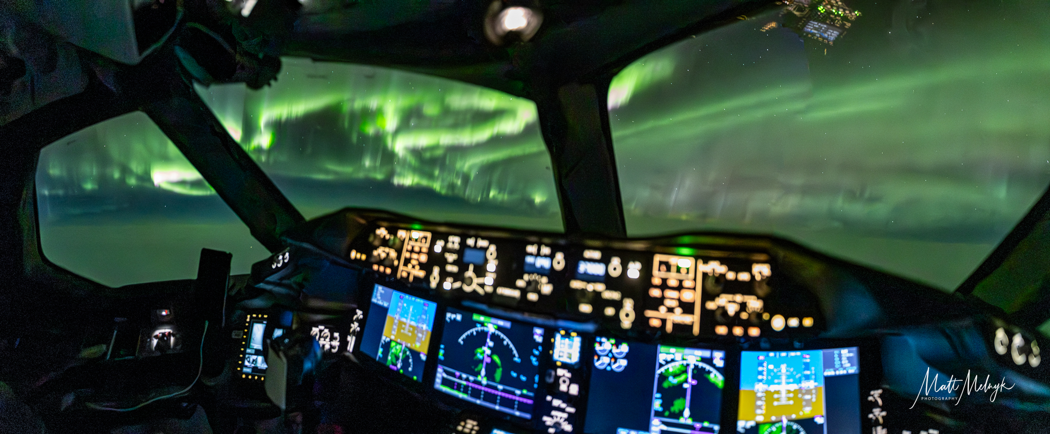 the view from inside the cockpit shows the controls of a plane and a strong aurora show with glowing ribbons of light as far as the eye can see.