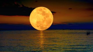 Full Moon June 2023: Orange color full moon in the night dark sky with light clouds above the sea with boats , copy space - stock photo.