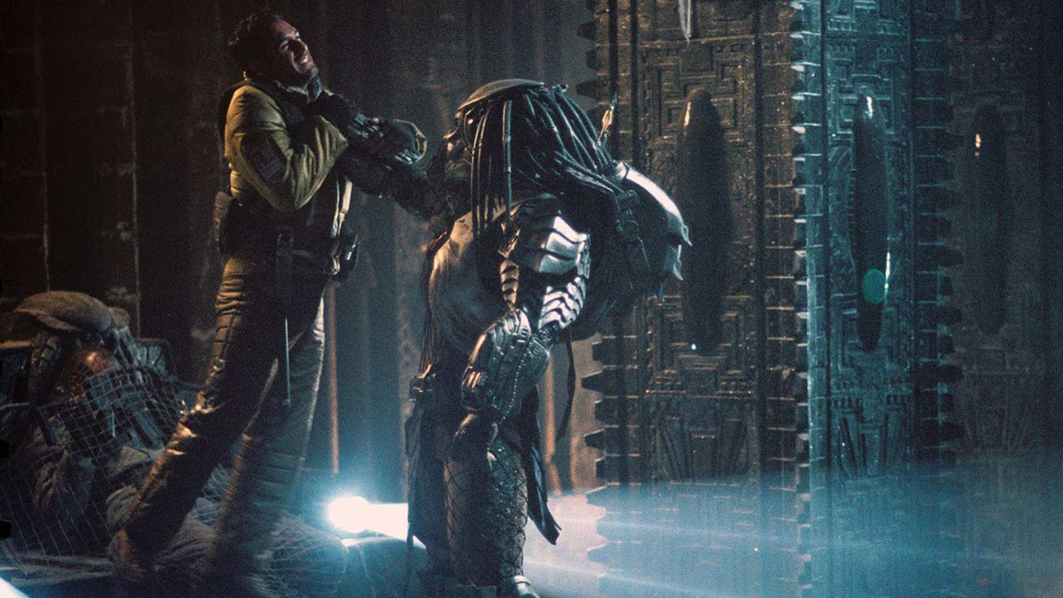 A still from the movie Alien Vs Predator in which a large Predator is holding up a character by the throat and there are ancient walls with inscriptions on them in the background.