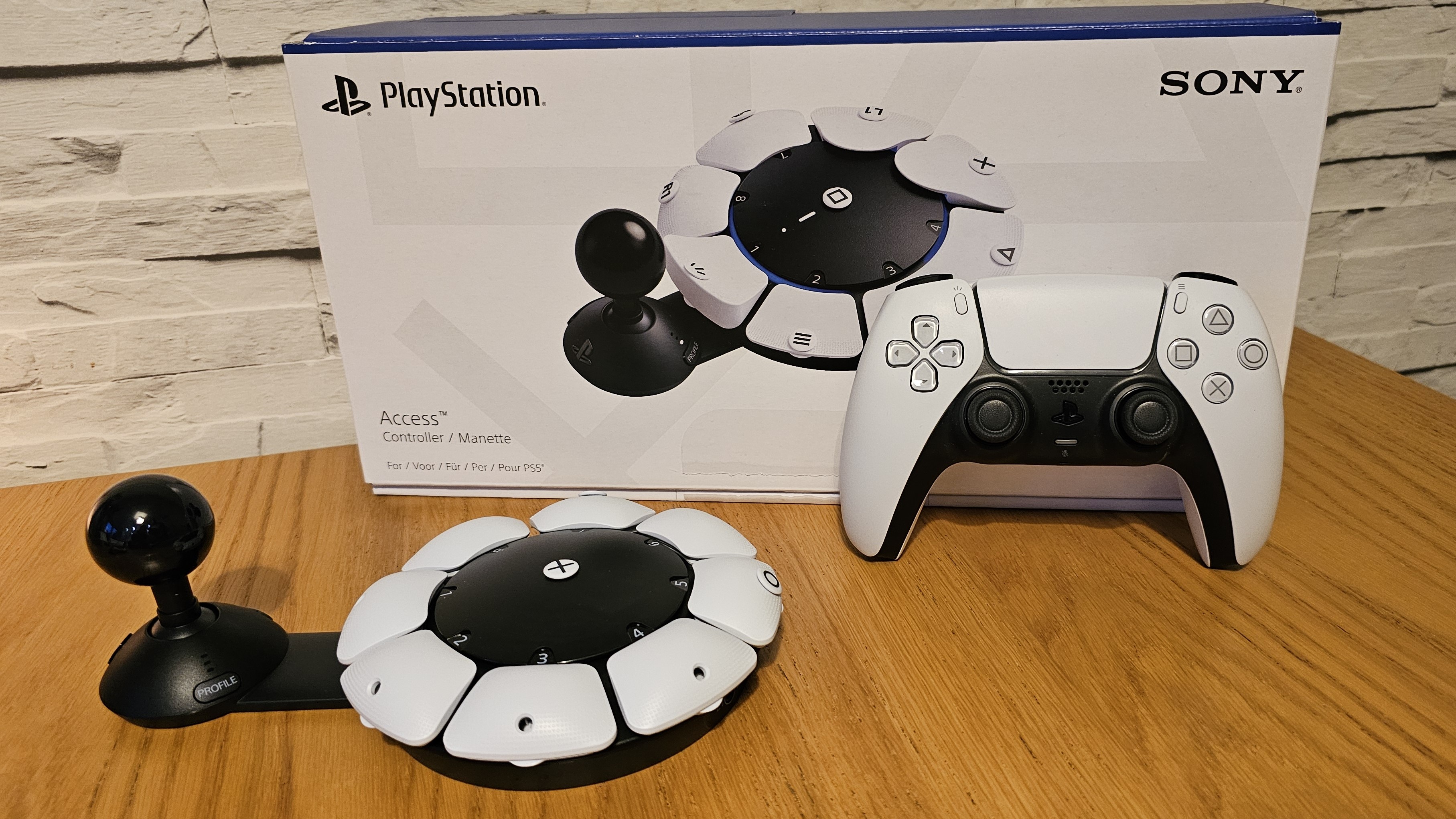 The PlayStation Access controller on a wooden surface next to its box and a PS5 DualSense controller