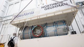 a white top half of a large shipping container is lifted by crane, exposing a metal cylindrical module inside.