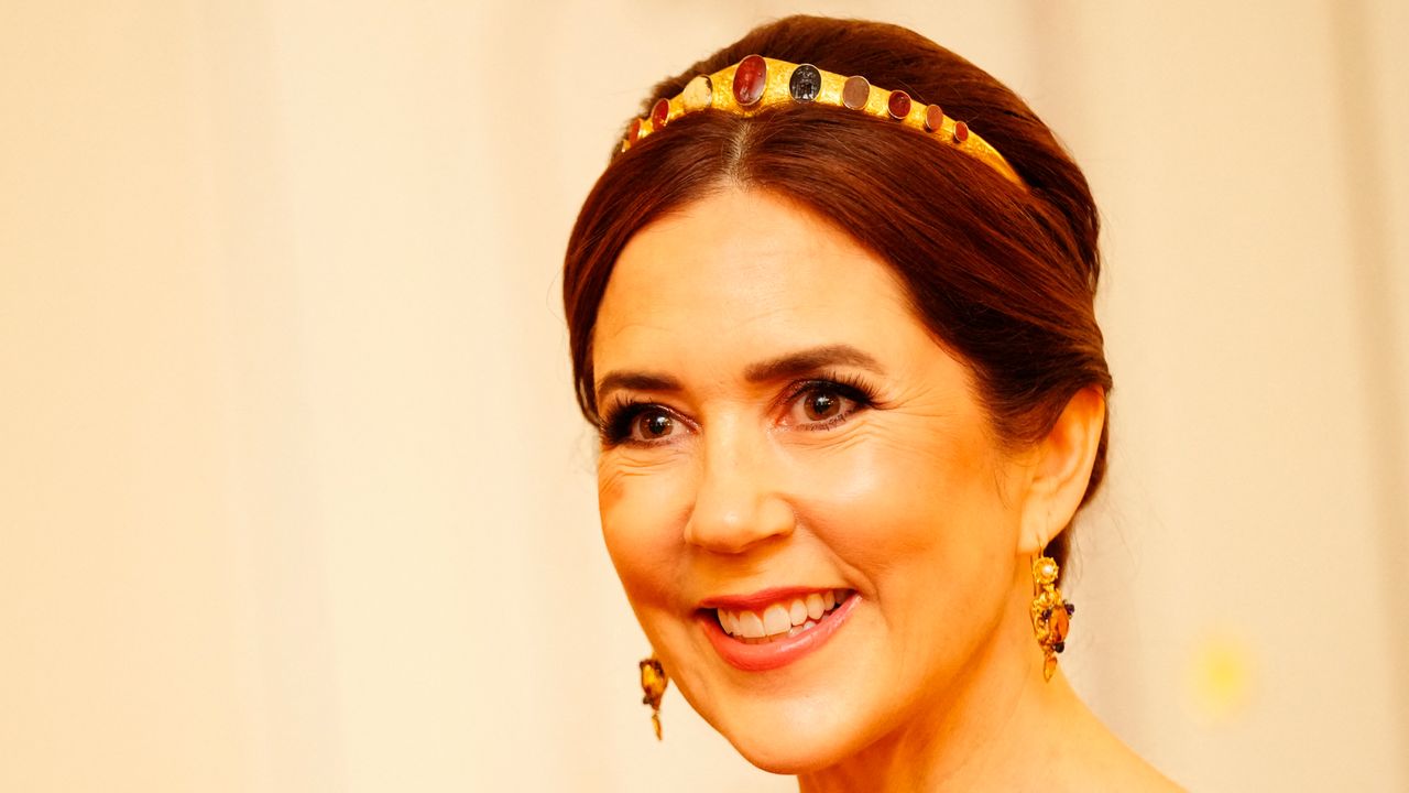 A headshot of Queen Mary wearing a slim gold tiara with stones on it and smiling in front of an ivory backdrop