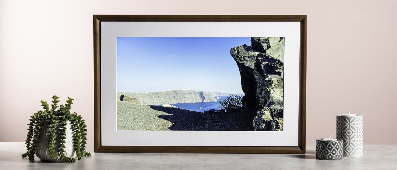 The Netgear Meural Canvas II showing a photo of rocky cliffs and an azure sea. It's surrounded by a plant and some patterned candle holders.