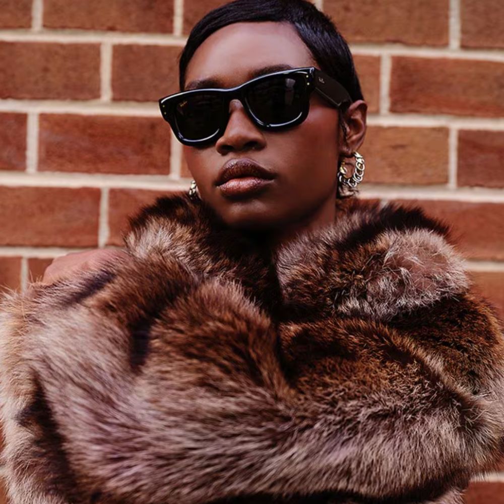 Young woman wearing Ray-Ban Wayfarer Puffer sunglasses and brown fake fur jacket, stood next to brick wall. 