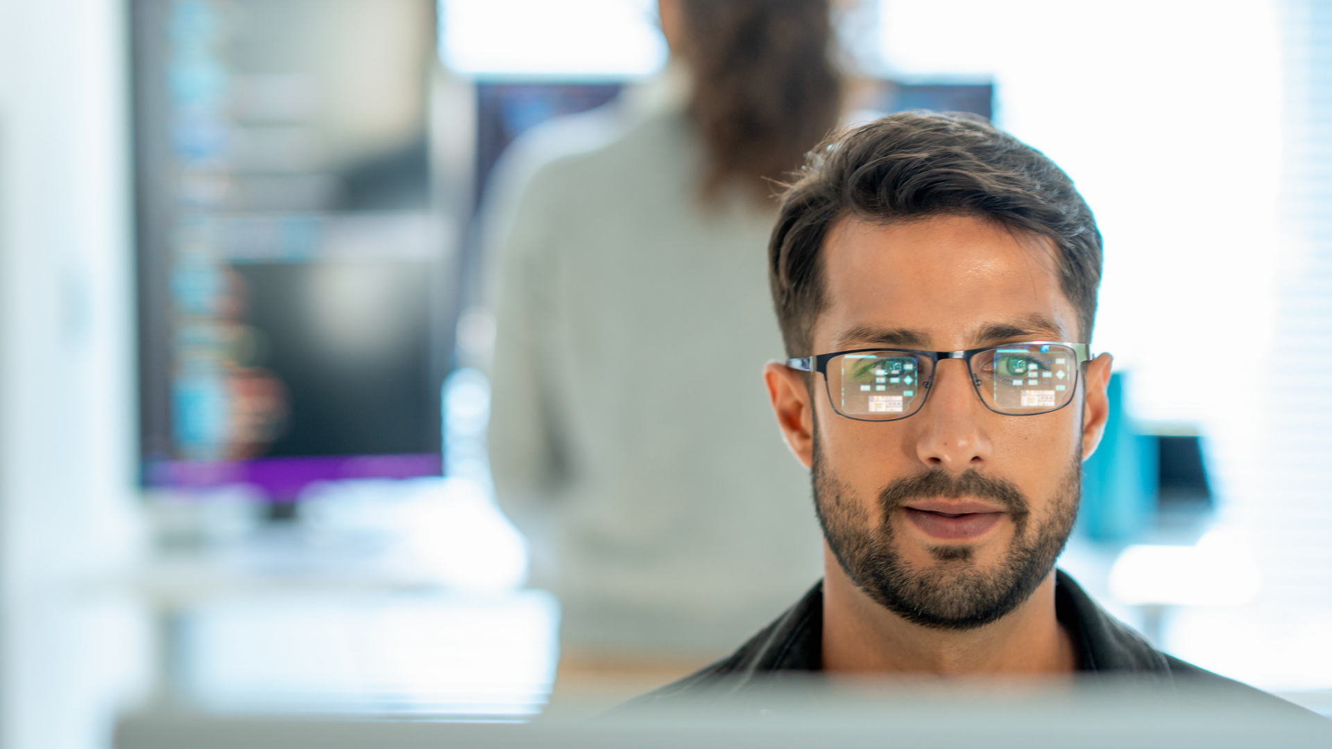 A man sitting in front of a computer in an office with the screen reflected in his glasses