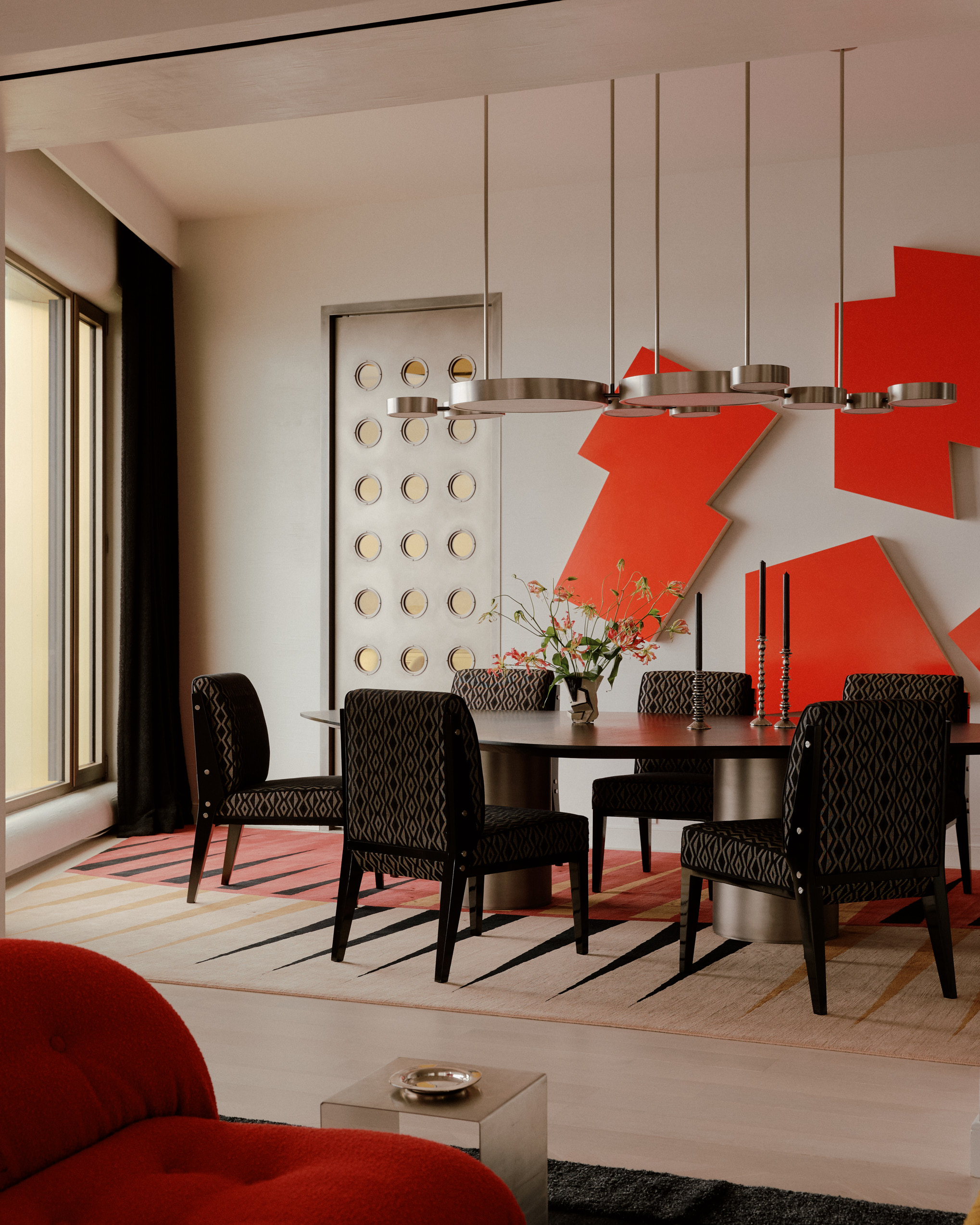 bold modern dining room with silver pendant light hanging over timber table with six dark upholstered chairs. a red sculpture hangs on the wall behind the table, and a door with holes in it leads through to the kitchen