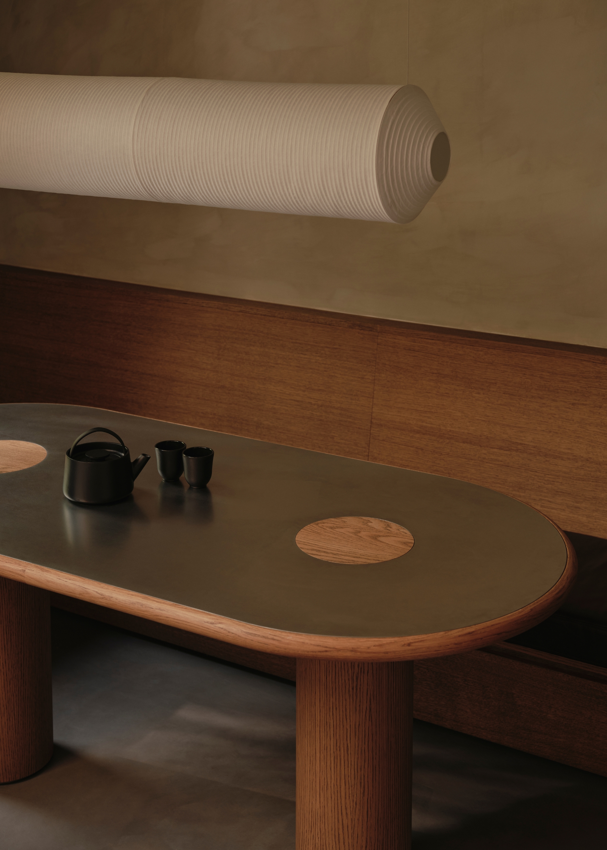 A calming dining room with a steel and wood curved dining table, a black teapot and glass set, a wood banquette seat, and limewashed walls beside a white cylindrical pendant light