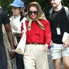 Naomi watts transforms into jackie kennedy wearing a headscarf, large sunglasses, a red blouse, brown belt, beige wide-leg pants, and brown woven leather ballet flats.