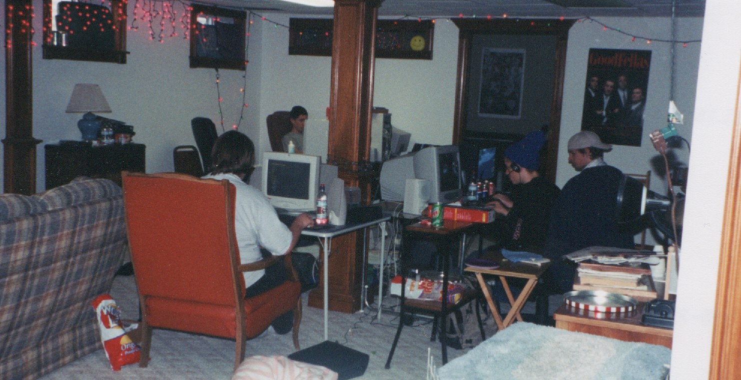 In a large living room, gamers sit at folding tables in rows, engrossed in games on CRT monitors.