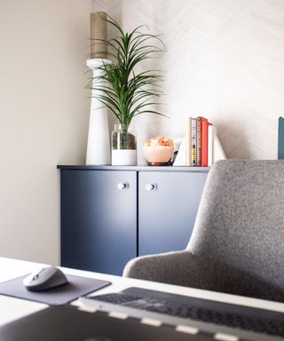A home office with a white desk with a black keyboard and mouse, gray office chair, onlooking a dark blue cabinet with a green spiked plant, orange bowl, and orange and brown books on it