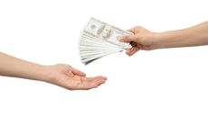 A hand holds out fanned-out hundred-dollar bills to another hand.