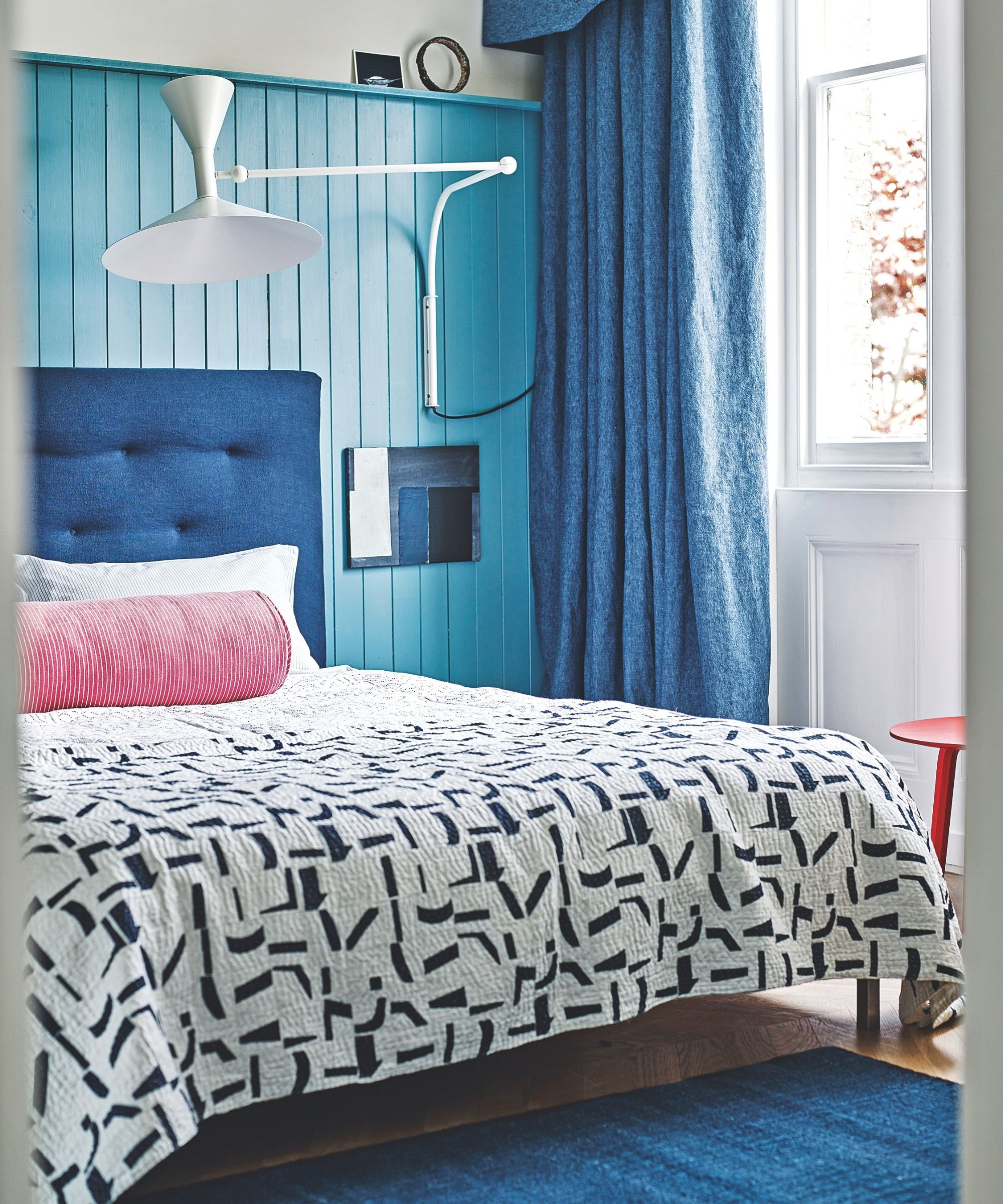 A blue panelled bedroom with a blue bed with patterned bedding and pink cushion, blue rug, white lamp, red stool, and large windows with long blue curtains