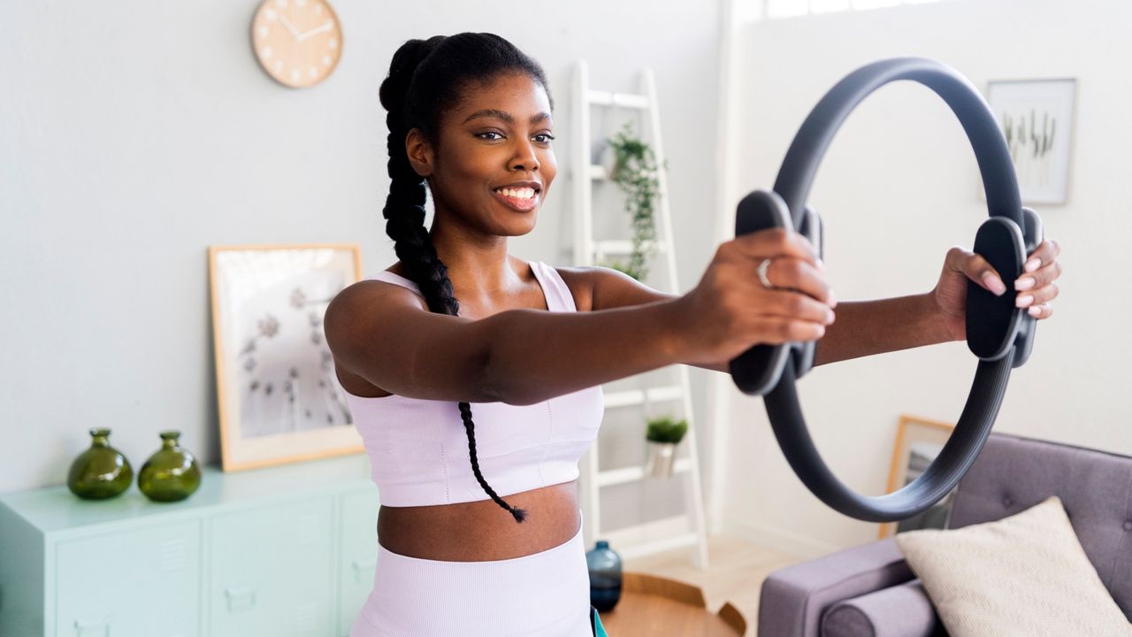 woman standing in a white living room space holding a pilates ring out in front of her