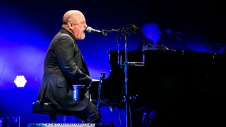 Billy Joel sits at his black piano with blue light behind him