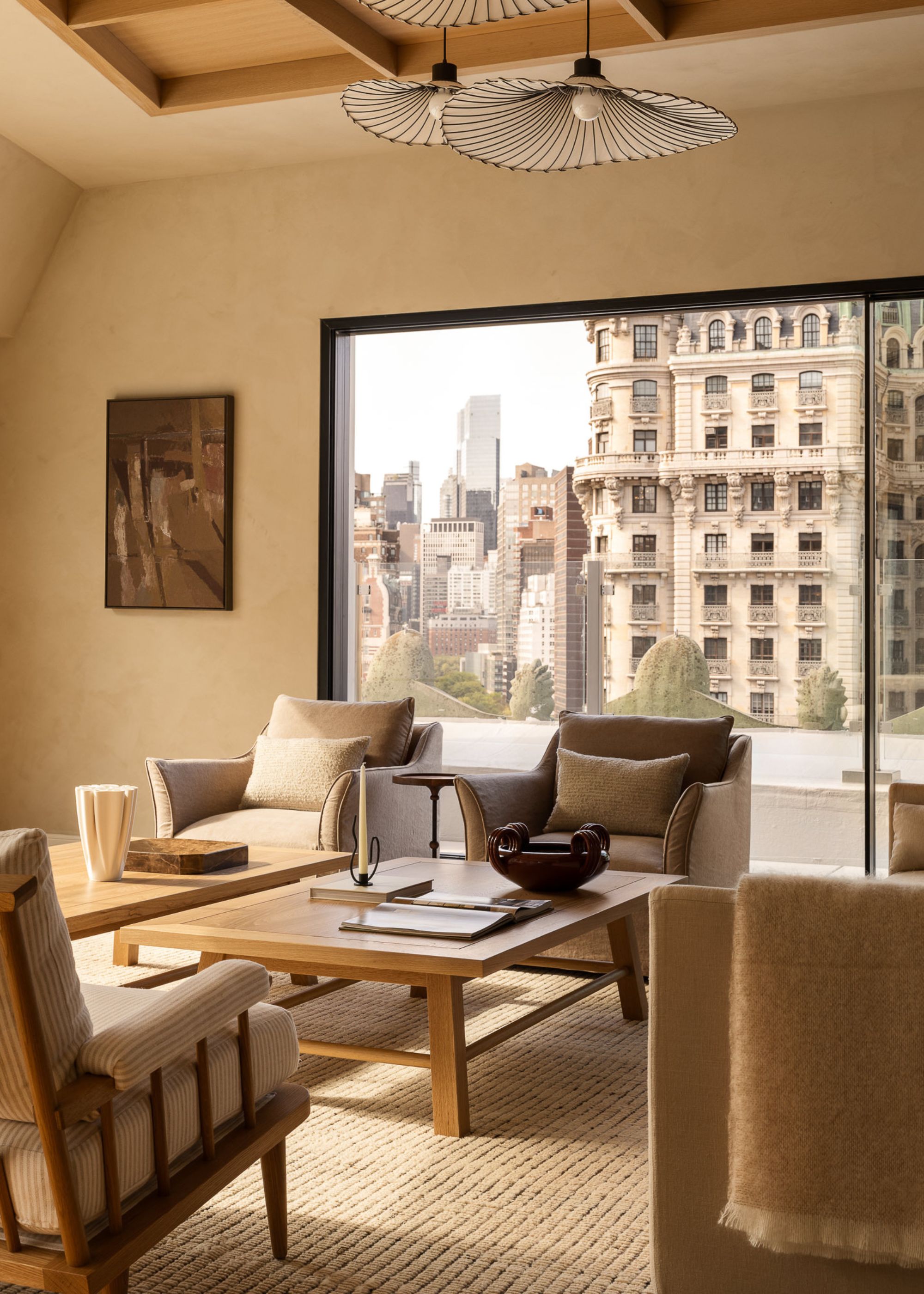A modern neutral living room in a penthouse with a city view from large sliding glass doors leading to a balcony with two wooden coffee tables and beige armchairs