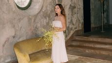 Kelly Wearstler stands in a brutalist room holding a bouquet of yellow flowers