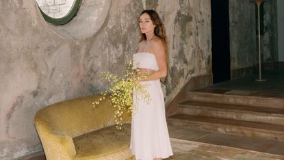 Kelly Wearstler stands in a brutalist room holding a bouquet of yellow flowers
