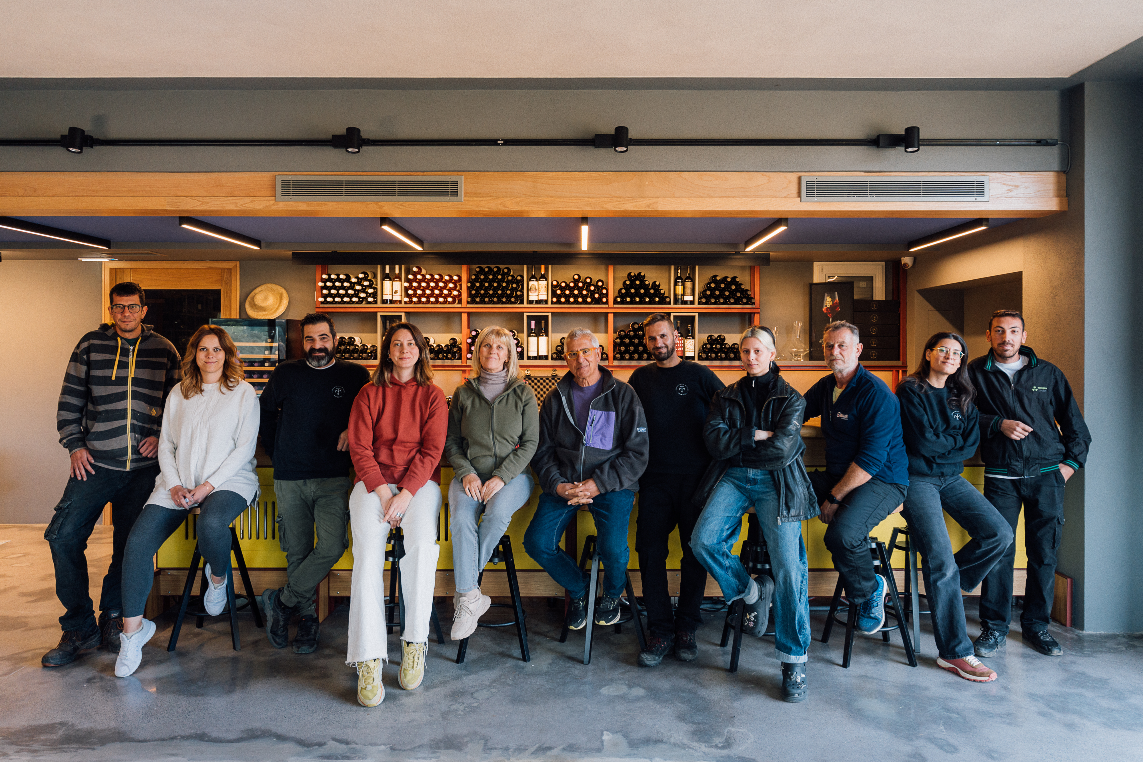The Tsimbidi family and winemaking team: 11 people sat on stools in front of a bar, with wine bottles in the background