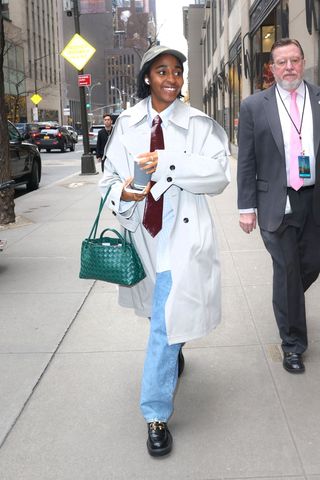 Ayo Edebiri wearing a grey trench coat with blue jeans, a leather tie, a green handbag, and a baseball cap