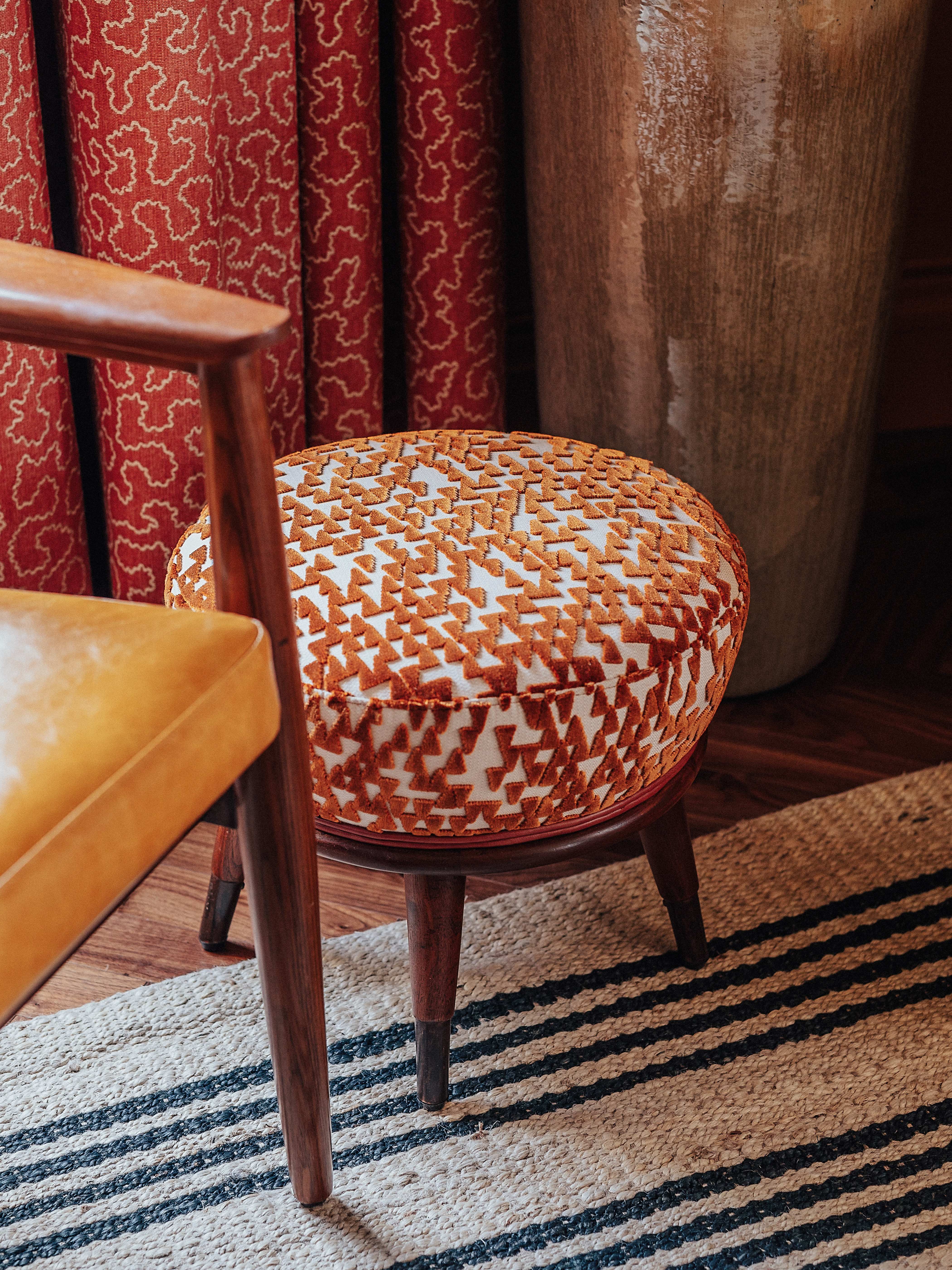 A layered, transitional hotel suite featuring a series of figurative, colorful artworks on its walls, patterned furniture, including rugs, armchairs, and sofas, split-color pillars, and a buttery, tile blue, and green palette.