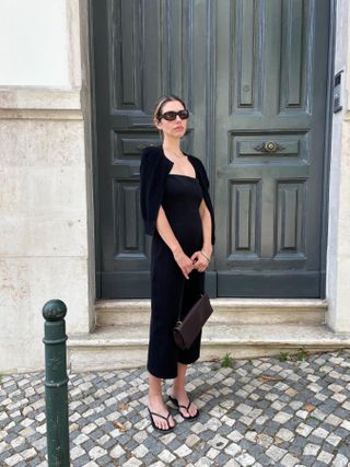 Robin Reetz on a street in Europe wearing a black dress with black flip-flop sandals, and a black cardigan over her shoulders.