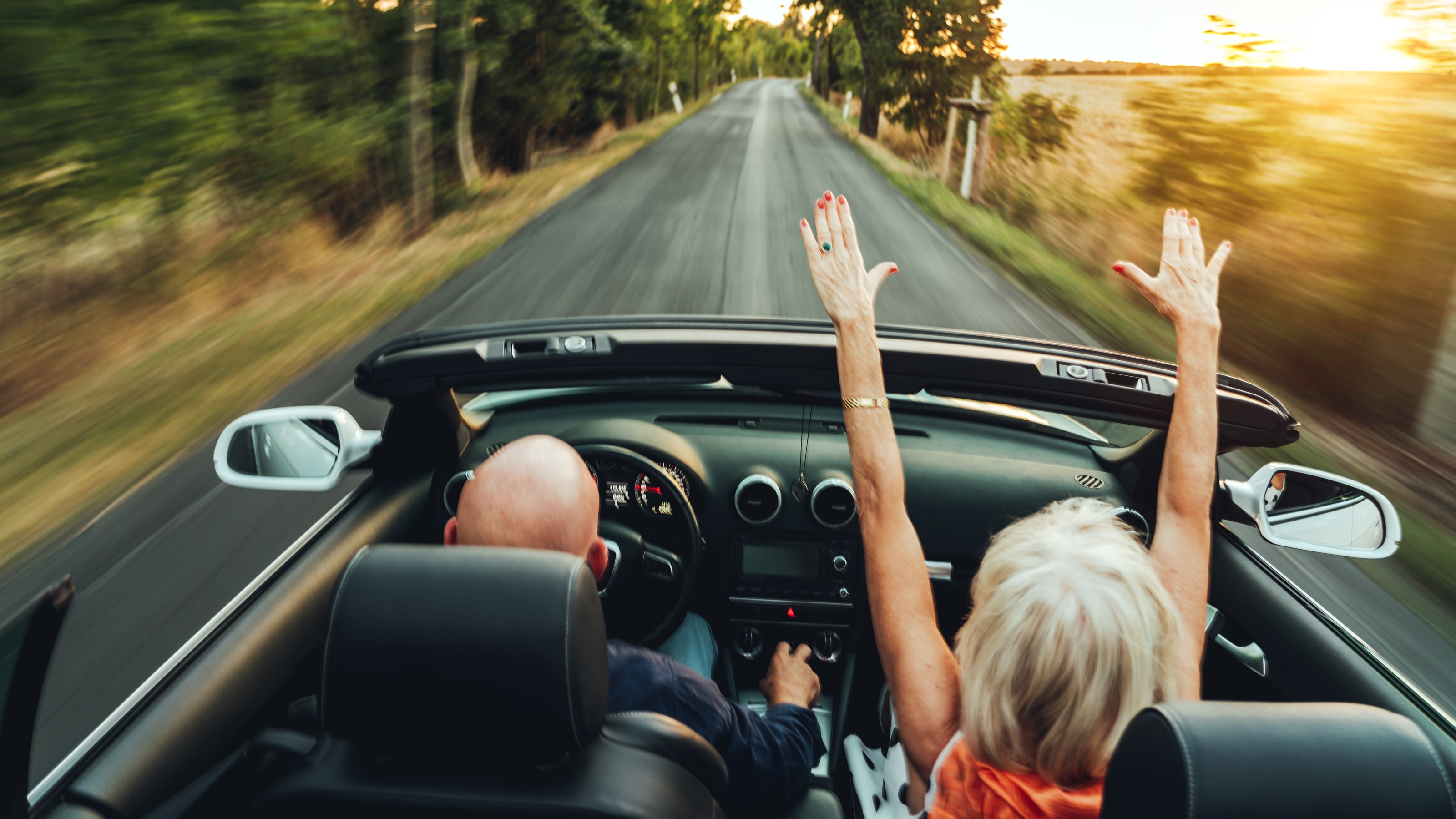 Rear view of an older couple driving down a country road in a convertible. 