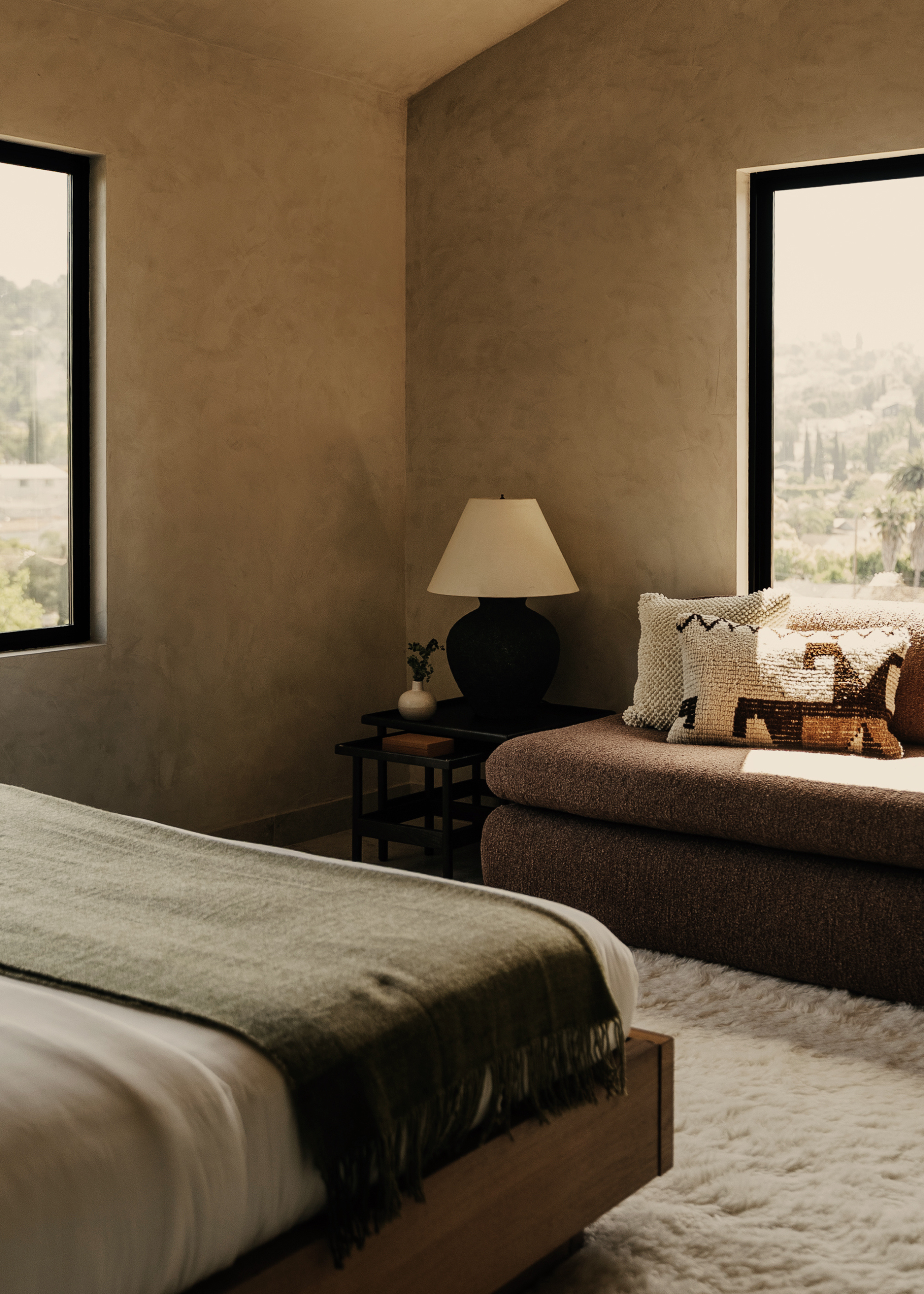 A bedroom with sandy limewash walls, side table with a lamp and a vase, a brown sofa bench with patterned pillows, and a fuzzy rug below a bed