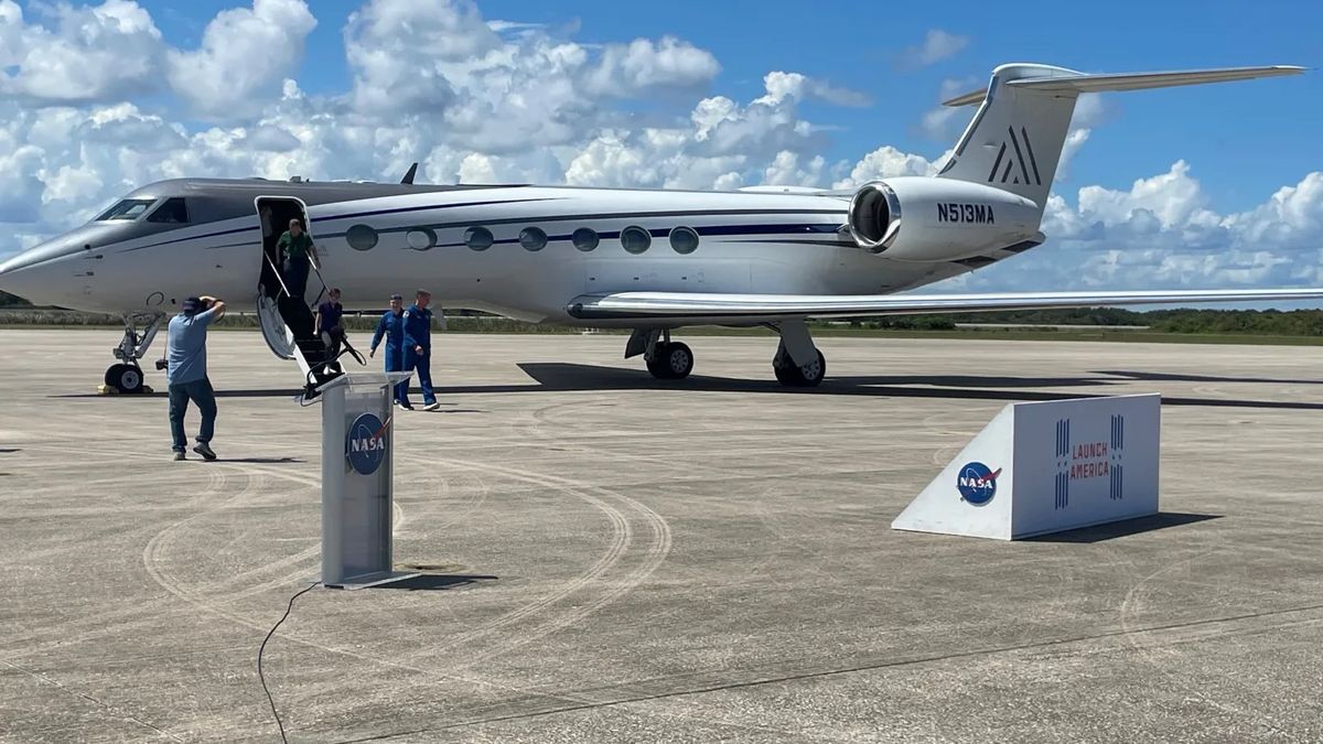 two people walk from a small white jet onto the tarmac, heading toward a lectern with the nasa emblem on it