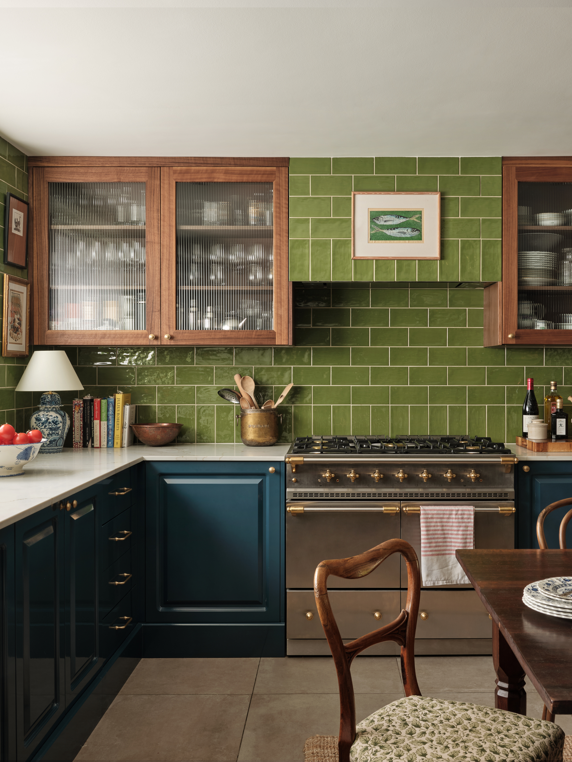 green kitchen with bright green tiled backsplash, timber upper cabinets with glass fronts, white counter, navy blue cabinetry, and gray floor tiles, with silver cooker and lamps and book styled on the counter, with a dining table and chairs in the middle of the room