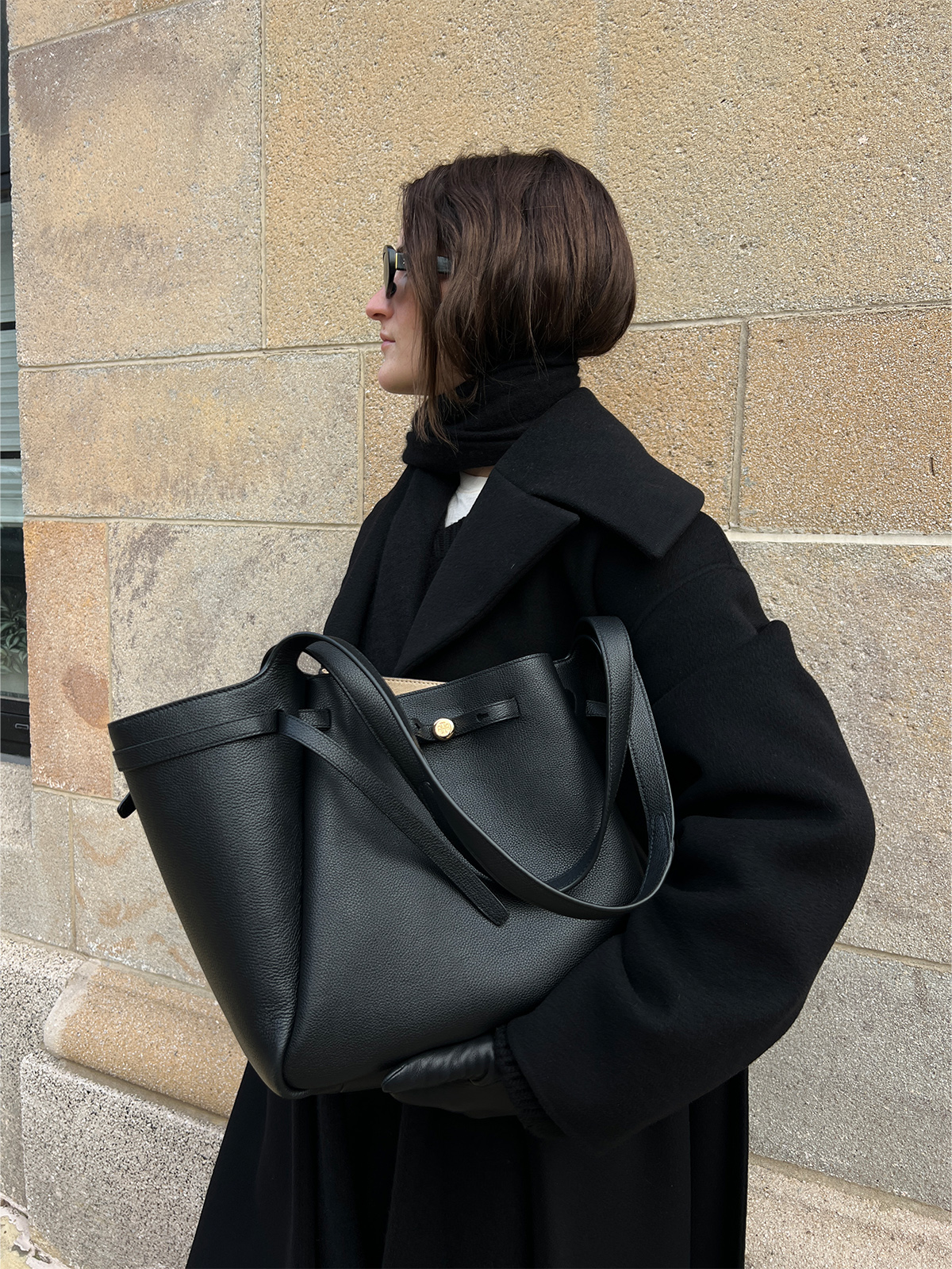 Eliza Huber testing out the Tory Burch Romy Tote Bag in black with a long black Nanushka coat, jeans, and a scarf.