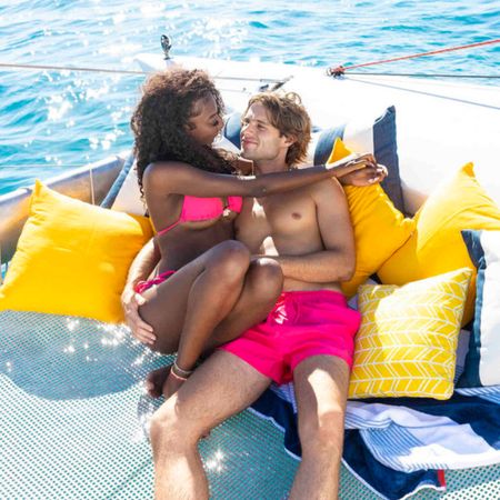 Trinity Tatum and Bryce Dettloff cuddle while sitting on a pile of yellow and white-and-blue-striped pillows on a boat on the Fijan ocean, in 'Love Island USA' season 8.
