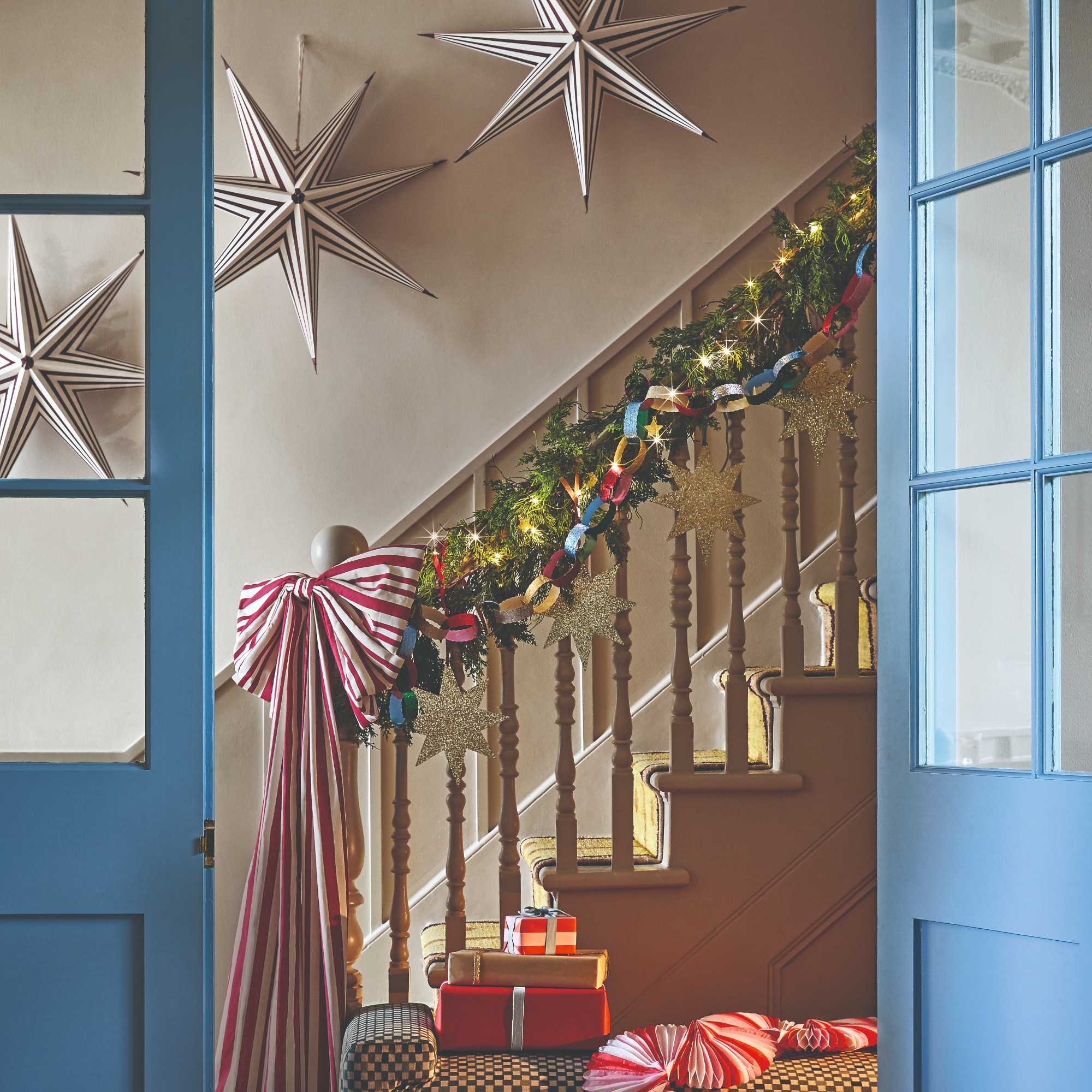 A staircase decorated for Christmas with an extra large striped bow, a garland and three striped paper Christmas decorations on the wall