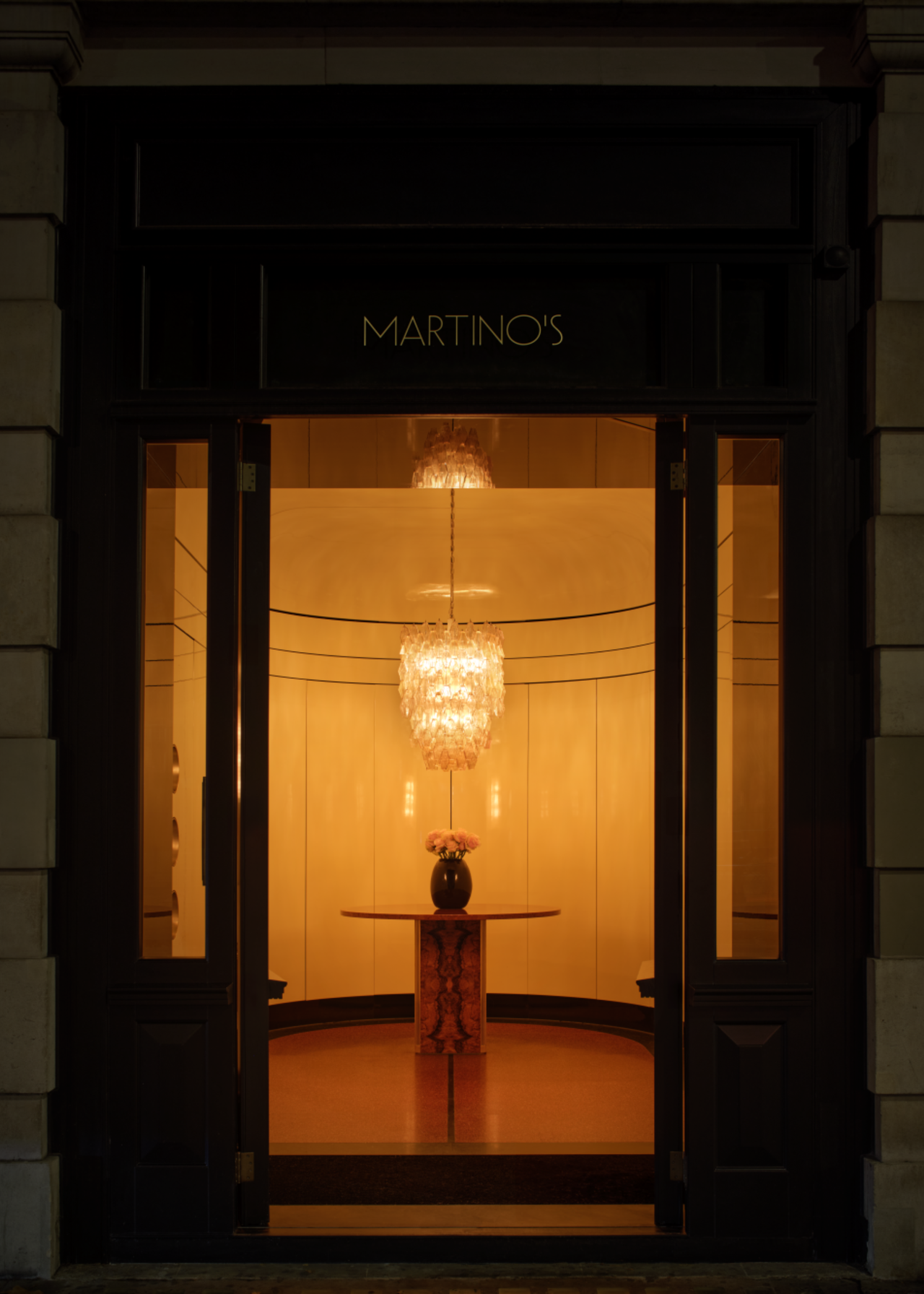 the entrance to martinos, a large art deco chandelier hangs over a small wooden table, in a curved, white room