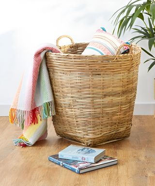 Large basket with colorful blanket draped over the edge and pillow inside beside plant on wood floor from A Place for Everything