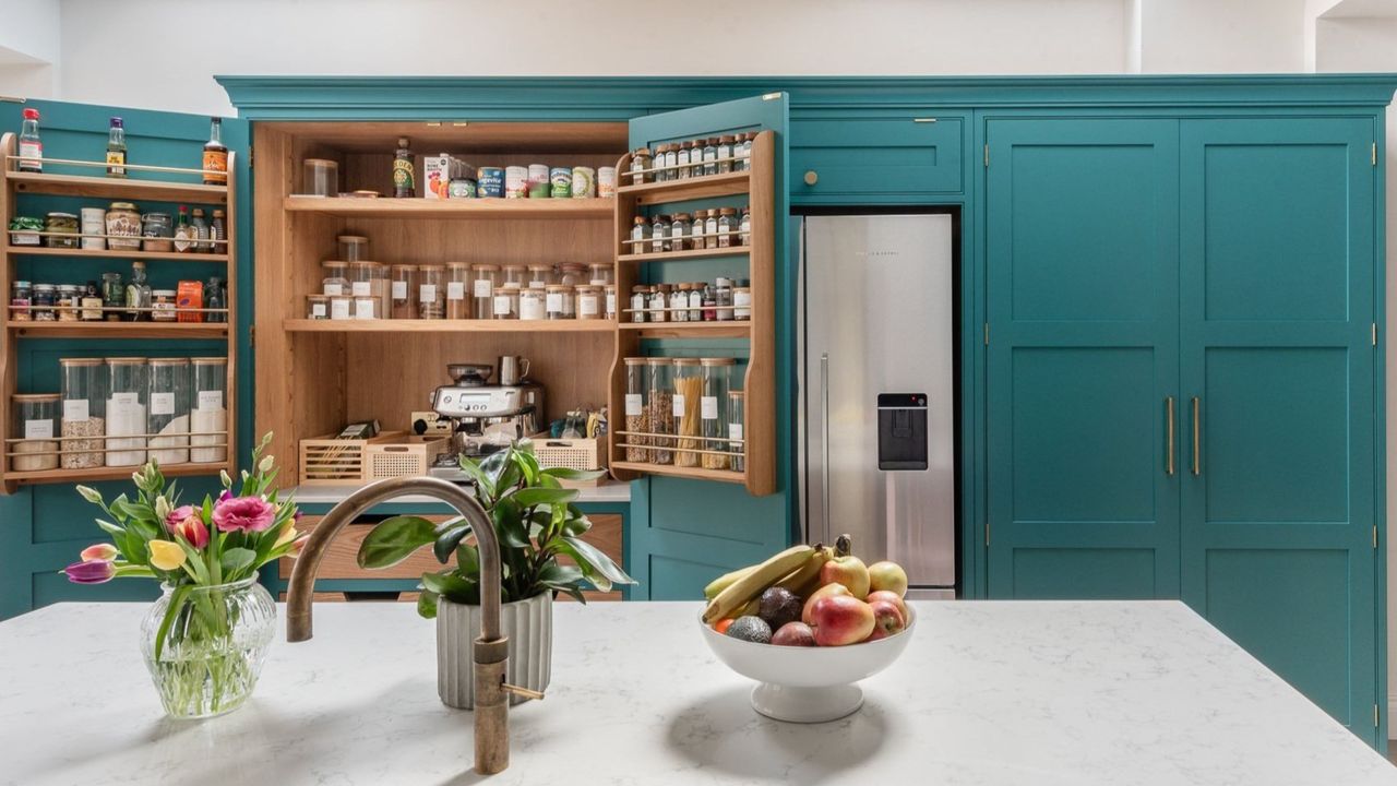 Ocean blue kitchen cabinets with a pantry and hidden storage. There is also a large island with a bowl of fruit on top as well as a plant pot and flowers.