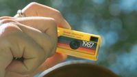 A hand holding up a yellow coloured Kodak Charmera with the sky and a tree behind him