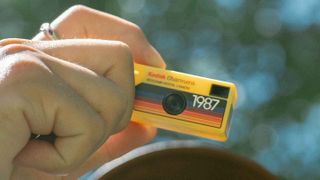 A hand holding up a yellow coloured Kodak Charmera with the sky and a tree behind him